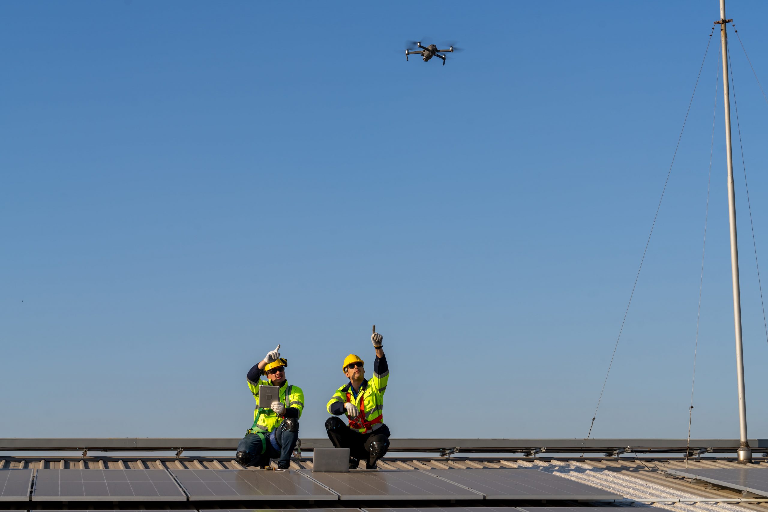 Zwei Ingenieure sitzen auf einem Dach mit PV-Anlage und lassen eine Drohne darüber fliegen