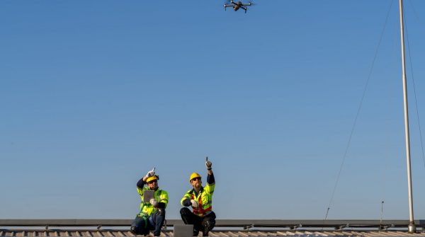 Zwei Ingenieure sitzen auf einem Dach mit PV-Anlage und lassen eine Drohne darüber fliegen